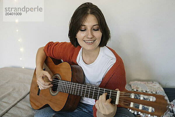 Smiling beautiful woman practicing guitar sitting in bedroom at home