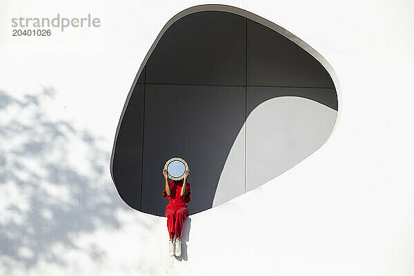 Woman in red jumpsuit with mirror sitting on white wall