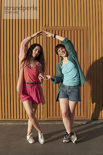 Smiling gay couple dancing together with holding hands in front of brown building wall