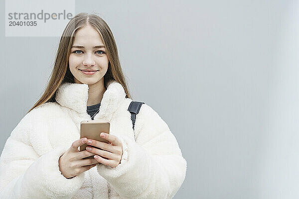 Smiling teenage girl using mobile phone in front of white wall