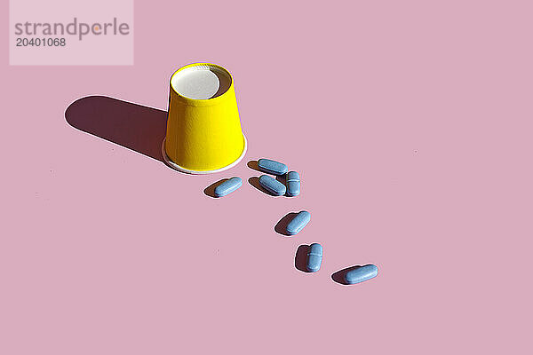 Yellow paper cup with blue pills over pink background