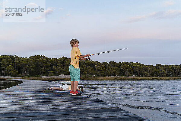 Brother with fishing rod standing by sister lying on pier over lake at sunrise