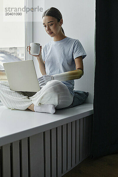 Young woman with prosthetic arm drinking coffee and using laptop on window sill at home