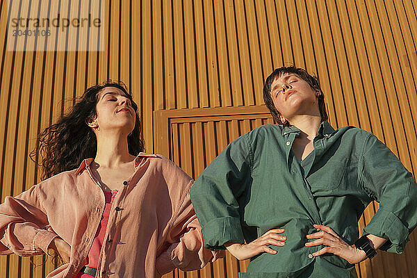 Gay women standing with arms akimbo in front of brown building wall