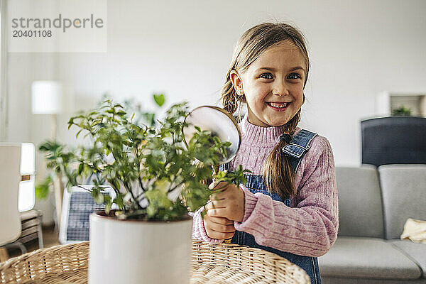 Smiling girl with magnifying glass by potted plant at home