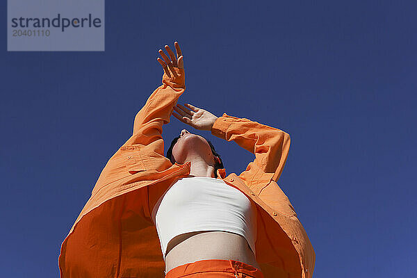 Woman in orange casuals dancing with arms raised under clear blue sky on sunny day