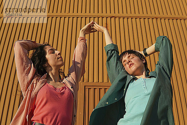 Hay couple with eyes closed holding hands in front of brown building wall