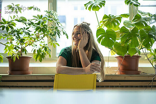 Smiling businesswoman sitting with hand on chin near potted plants in office