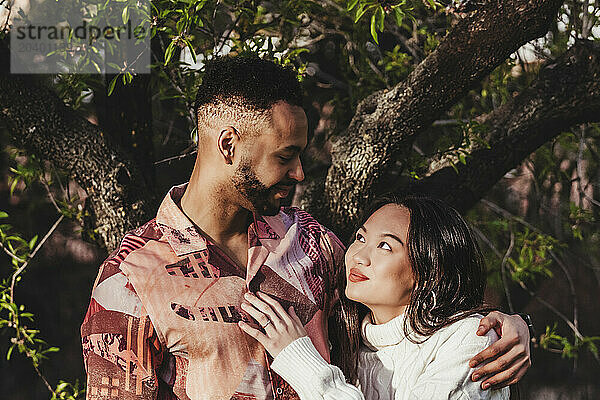 Smiling man embracing girlfriend near tree at park