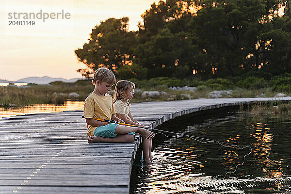 Siblings fishing together sitting on pier in lake at sunrise