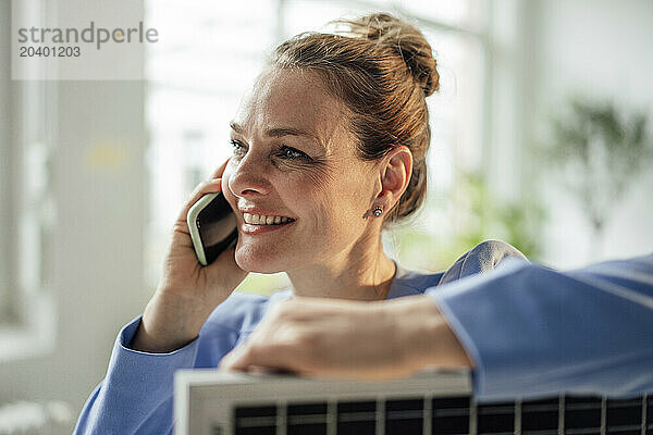 Smiling mature businesswoman talking on smart phone with solar panel in office