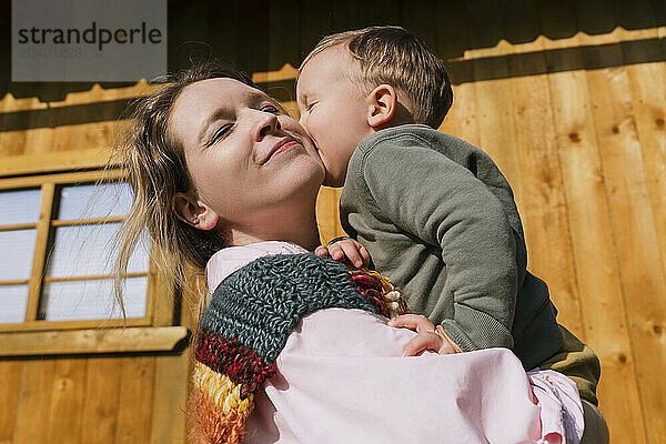 Boy kissing mother near house on sunny day