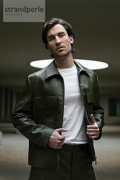 Fashionable young man wearing green leather jacket standing in illuminated parking garage