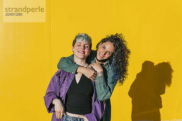 Happy woman embracing lesbian friend in front of yellow wall