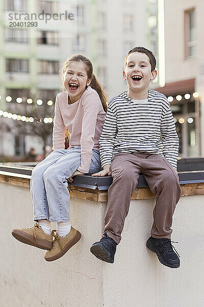 Cheerful boy sitting with sister on wall