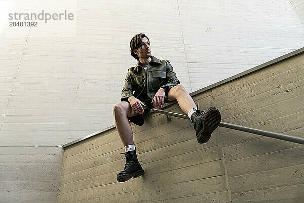 Fashionable young handsome man wearing green leather jacket and shorts sitting on wall