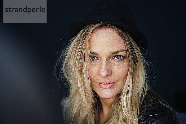 Confident mature blond woman wearing hat against black background