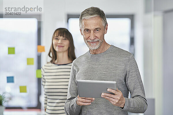 Confident businessman holding tablet PC with colleague in background