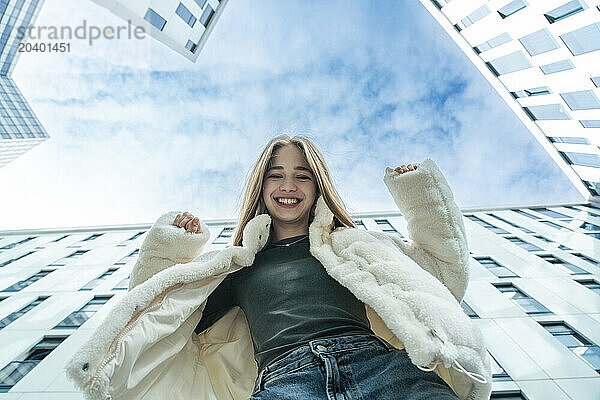 Happy teenage girl wearing white jacket standing near modern building