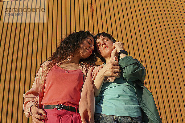 Gay couple standing with eyes closed in front of brown building wall