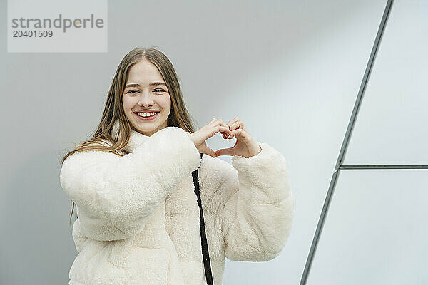 Smiling teenage girl showing heart sign in front of white wall