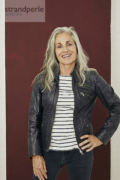 Happy woman in leather jacket standing with hand on hip against maroon background