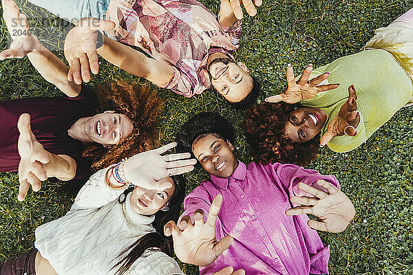 Smiling multiracial friends lying down on grass with hands raised at park