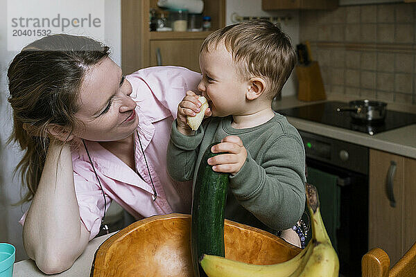 Smiling mother looking at boy eating cucumber in kitchen