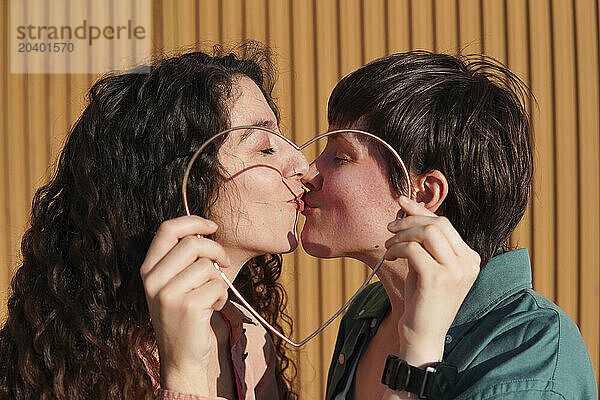 Lesbian couple kissing with metal heart shape in front on sunny day