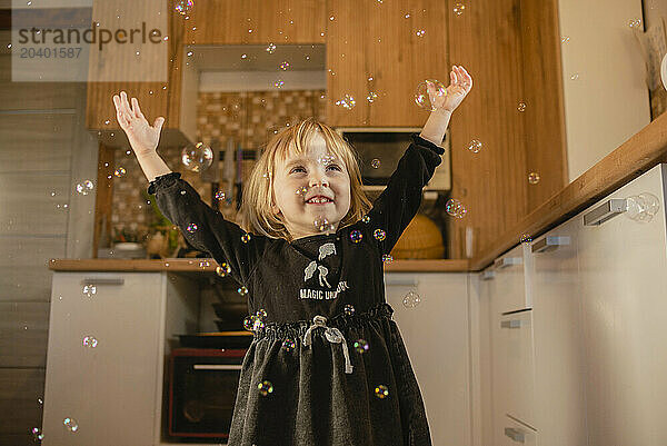 Smiling girl with arms raised playing with bubbles in kitchen at home