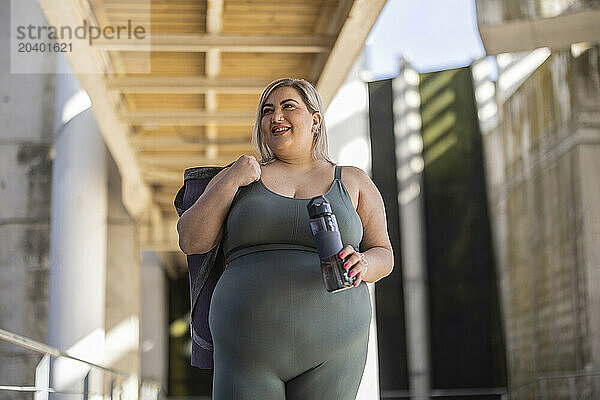 Happy young curvy woman standing with exercise mat and water bottle