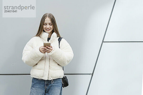 Smiling teenage girl using smart phone standing in front of white wall