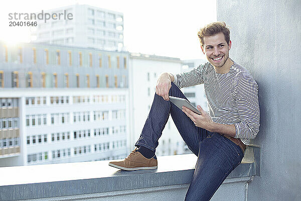 Happy young man sitting on wall with tablet PC in balcony