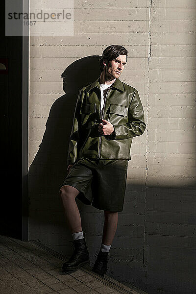 Fashionable young handsome man wearing green leather jacket and shorts leaning on wall