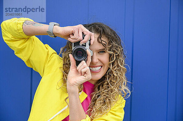 Happy woman photographing through camera in front of blue wall