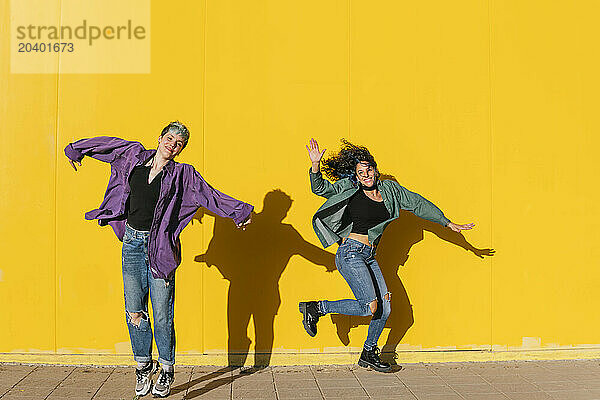 Happy lesbian couple jumping in front of yellow wall at sunny day