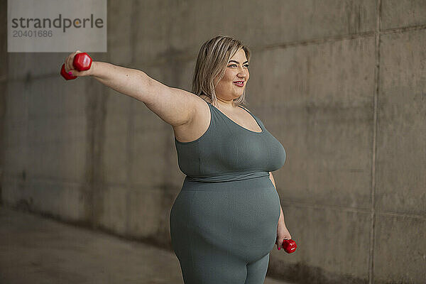 Smiling curvy woman exercising with dumbbells near wall