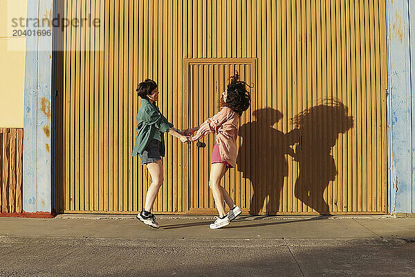 Cheerful gay couple dancing together with holding hands in front of brown building wall