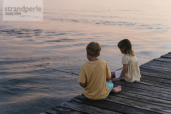 Brother and sister fishing together sitting on pier in lake at sunrise