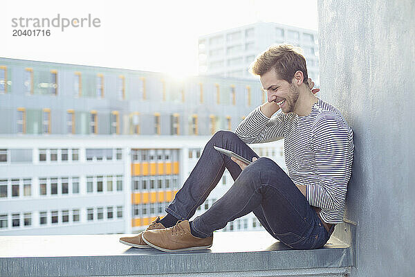 Happy young man using tablet PC on wall in front of buildings