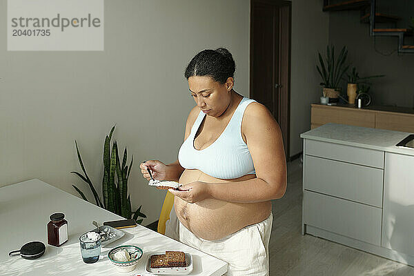 Pregnant woman spreading cream cheese on bread for breakfast at home