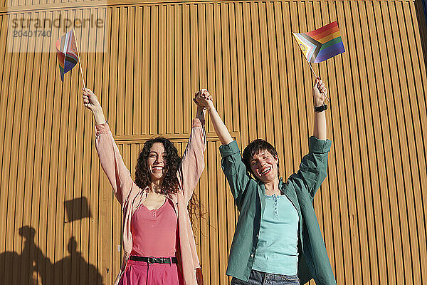 Happy lesbian couple holding hands and raising LGBTQIA pride flag in front of brown building wall