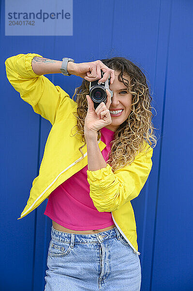 Smiling woman photographing through camera in front of blue wall