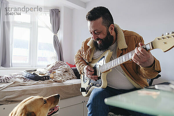 Smiling man playing guitar sitting next to dog at home