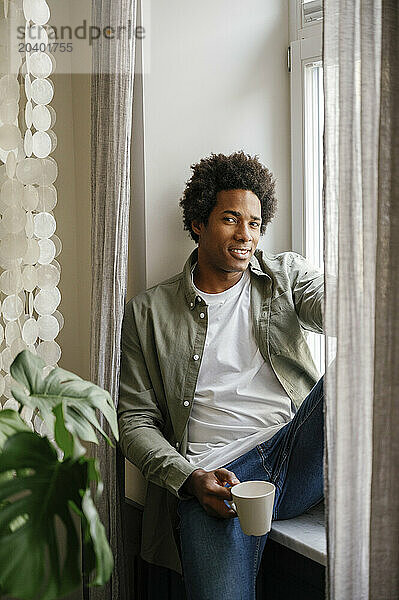 Smiling man with coffee cup sitting on window sill at home