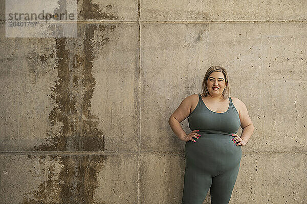 Smiling curvy woman standing with hands on hip in front of wall