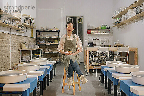 Confident craftsperson sitting on stool amidst pottery wheels at workplace