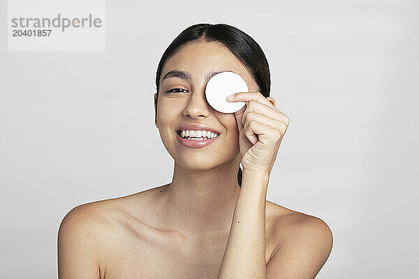 Smiling beautiful young naked woman covering eye with cotton pad against white background