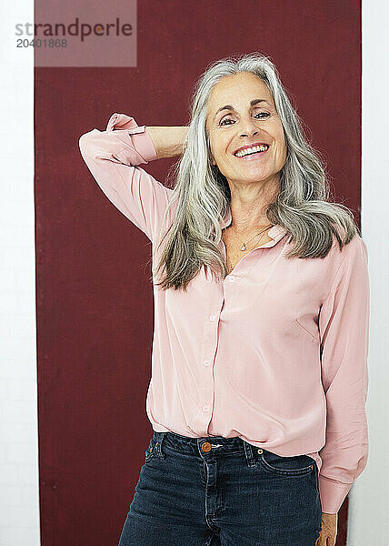 Smiling woman with hand behind head standing against maroon background