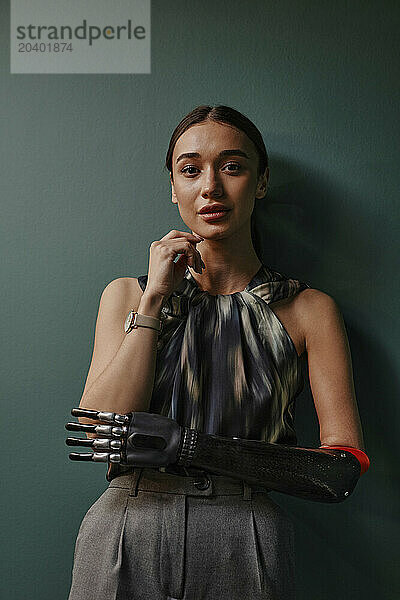 Confident woman with black bionic arm leaning on green wall